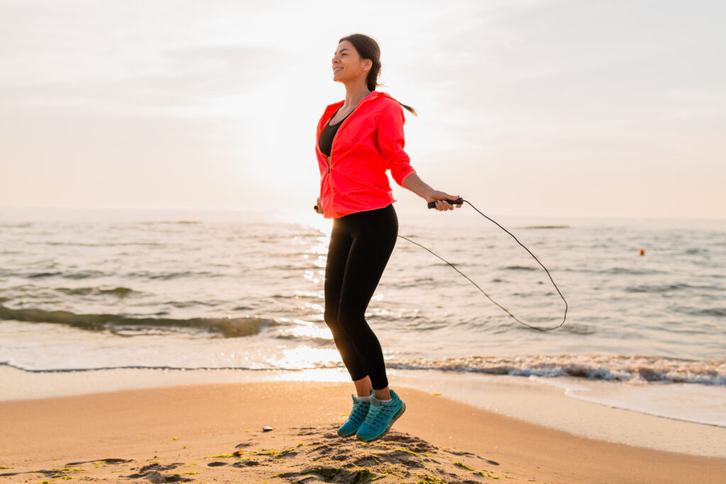 young attractive woman doing sport exercises in morning sunrise on sea beach in sports wear, healthy lifestyle, listening to music on earphones, wearing pink windbreaker jacket, jumping in jump rope